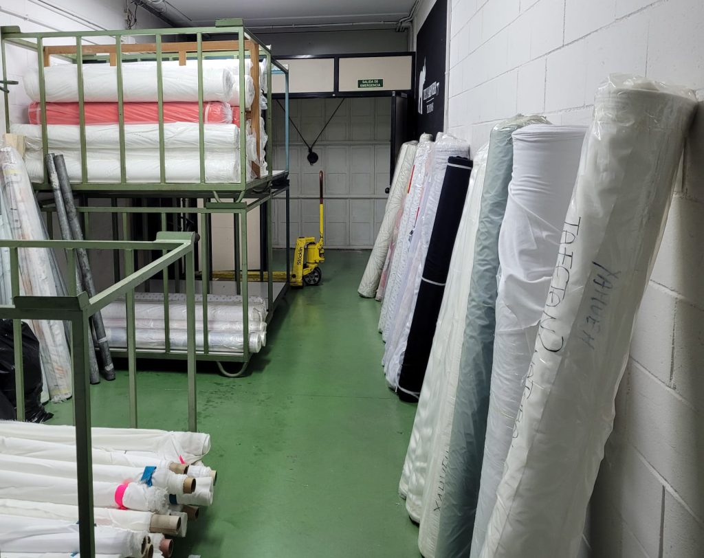 Vista del almacén de Texyahveh con rollos de tela protegidos y paletas de envío, junto a un transpalet manual, destacando la eficiencia en la logística del material